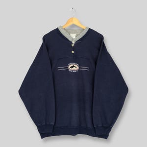 Puede incluir: Sudadera azul marino con cuello gris y tres botones. La sudadera presenta un gráfico de un delfín y el texto "Kona Harbor Hawaii, Porter Island, U.S." en el pecho. La prenda está colgada en una percha de madera.