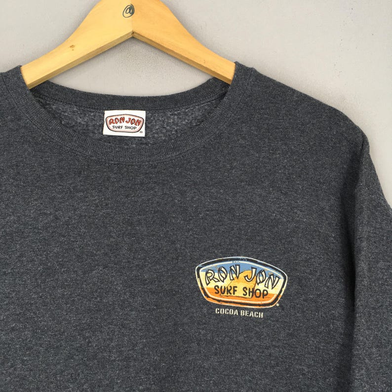 May include: A dark grey sweatshirt featuring the "Ron Jon Surf Shop" logo on the chest and lower right. The logo is in blue, yellow, and orange. The sweatshirt is displayed on a wooden hanger.