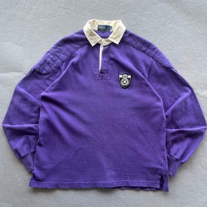May include: A long-sleeve purple rugby shirt featuring a cream-colored collar, a button-up placket, and ribbed shoulder panels. A black and white shield-shaped patch with the text Polo is embroidered on the left chest, and a Polo label is at the neck.