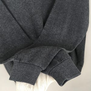 May include: Close-up of a dark grey sweatshirt sleeve with a ribbed cuff. The fabric has a heathered texture. A portion of a white knitted garment is visible beneath the sleeve.