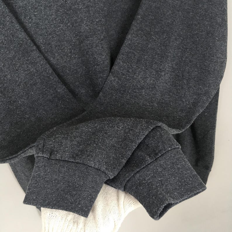 May include: Close-up of a dark grey sweatshirt sleeve with a ribbed cuff. The fabric has a heathered texture. A portion of a white knitted garment is visible beneath the sleeve.