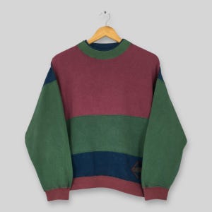 May include: A color-blocked sweatshirt in burgundy, green, and navy blue, with a diamond-shaped emblem on the lower right. The long-sleeved top has a crew neck and is hanging on a wooden hanger.