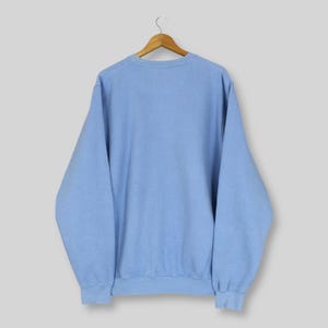 Puede incluir: Una sudadera azul claro cuelga de una percha de madera. La sudadera de cuello redondo tiene mangas largas y un dobladillo y pu&ntilde;os acanalados. La prenda es de color liso y parece estar hecha de un material suave.