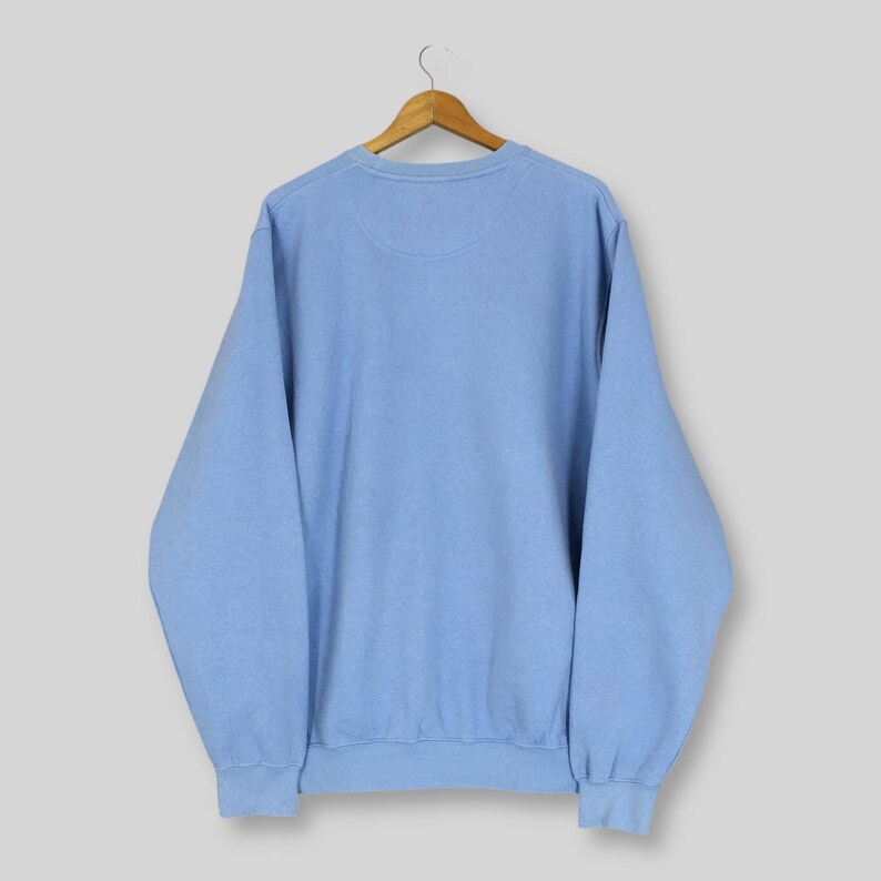 Puede incluir: Una sudadera azul claro cuelga de una percha de madera. La sudadera de cuello redondo tiene mangas largas y un dobladillo y pu&ntilde;os acanalados. La prenda es de color liso y parece estar hecha de un material suave.