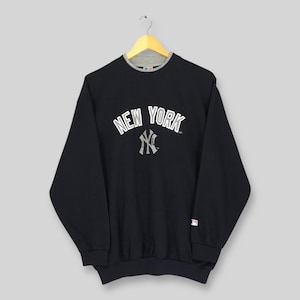 Può includere: Una felpa nera con la scritta bianca "NEW YORK" ad arco e un logo "NY" grigio. La felpa ha un colletto grigio e un piccolo logo sulla manica.
