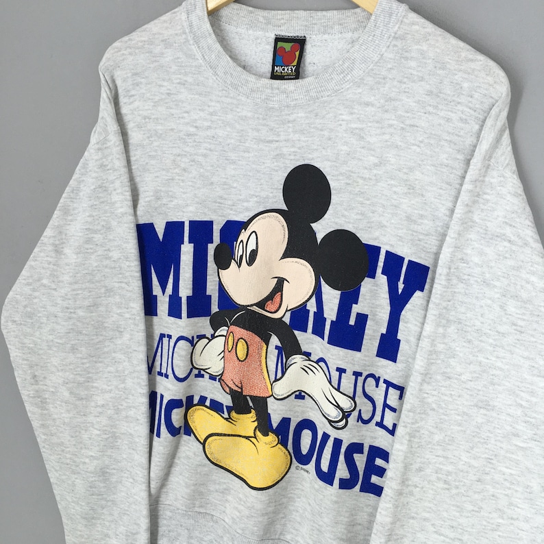 K&ouml;nnte beinhalten: Ein graues Mickey Mouse Sweatshirt mit einer gro&szlig;en Grafik von Mickey Mouse und den Worten "Mickey Mouse" in Blau. Das Sweatshirt hat einen Rundhalsausschnitt und lange &Auml;rmel. Das Mickey Mouse Logo befindet sich auf dem Etikett.