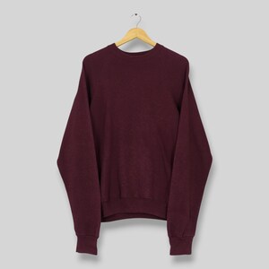 May include: A solid dark maroon long-sleeve sweatshirt featuring a classic crew neckline, raglan sleeves, and ribbed cuffs at the wrists and hem. The garment is displayed on a wooden hanger, showcasing a simple, plain design with no patterns or text.