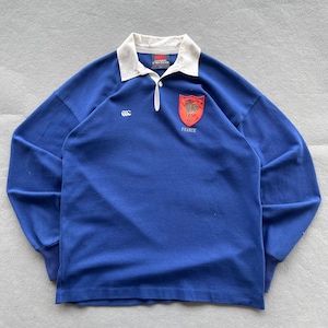 May include: A long-sleeve blue rugby shirt with a white collar and a button placket. The chest features a white Canterbury logo on the right and a red shield patch with a gold rooster emblem and the word FRANCE embroidered in white on the left.