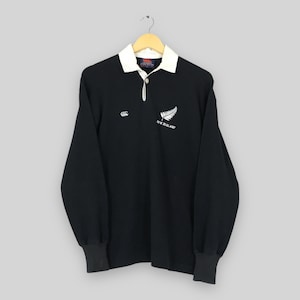 May include: Black long-sleeve rugby shirt with a white collar and cuffs. Features a white fern and "NEW ZEALAND" text on the chest. The shirt has a button-up collar and a small logo on the left chest.