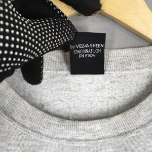 K&ouml;nnte beinhalten: Nahaufnahme eines grauen Sweatshirts mit Rundhalsausschnitt und einem schwarzen Etikett. Auf dem Etikett steht "by VELVA SHEEN CINCINNATI, OH RN 61626". Das Sweatshirt ist aus weichem, strukturiertem Stoff.