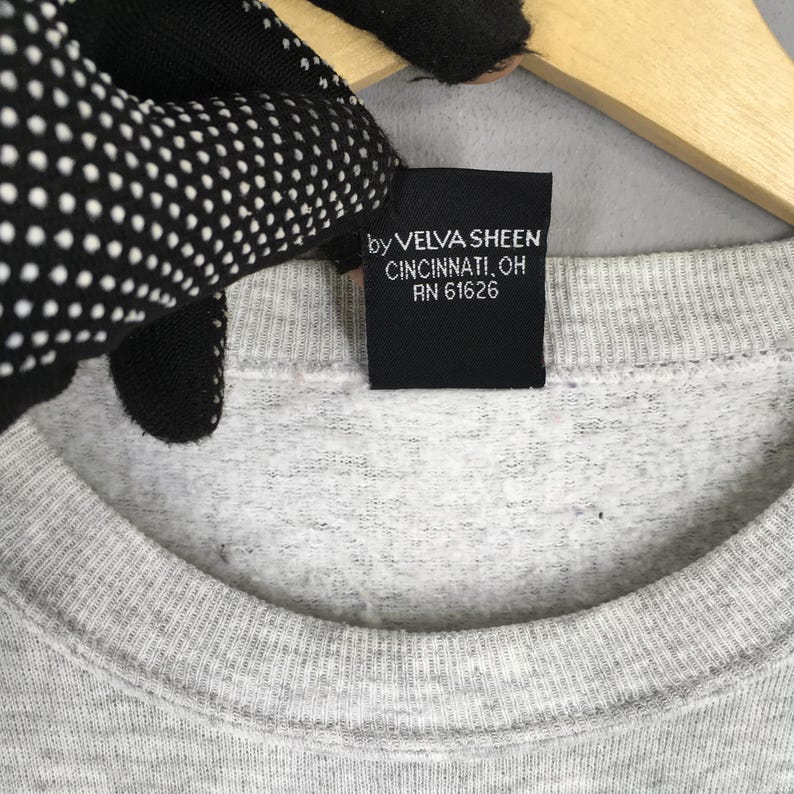K&ouml;nnte beinhalten: Nahaufnahme eines grauen Sweatshirts mit Rundhalsausschnitt und einem schwarzen Etikett. Auf dem Etikett steht "by VELVA SHEEN CINCINNATI, OH RN 61626". Das Sweatshirt ist aus weichem, strukturiertem Stoff.
