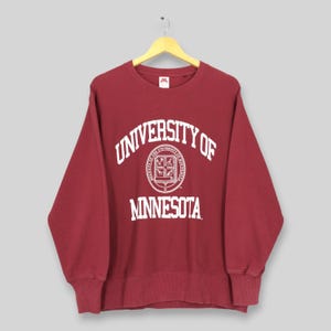 Puede incluir: Una sudadera de cuello redondo color granate con el texto blanco University of Minnesota arqueado sobre un sello universitario circular. La prenda tiene mangas largas con puños y dobladillo acanalados, y se observa una etiqueta blanca en el cuello.