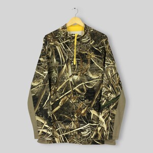 Puede incluir: Un jersey de camuflaje de manga larga con cremallera y cuello amarillos. La camisa presenta un estampado marr&oacute;n, verde y beige, con las palabras "Realtree Wetlands" bordadas en el pecho. Las mangas son de color beige liso.