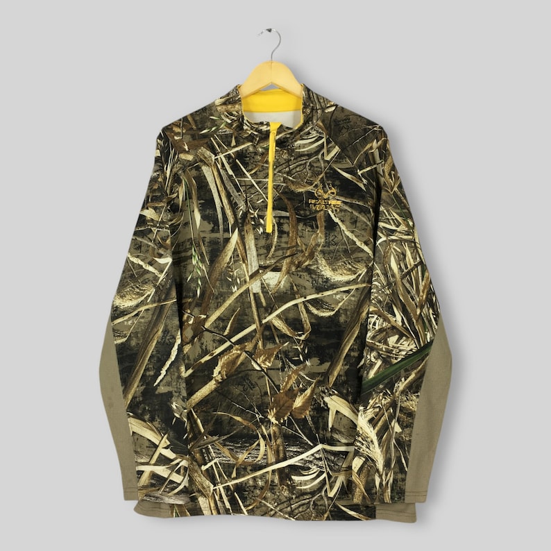 Puede incluir: Un jersey de camuflaje de manga larga con cremallera y cuello amarillos. La camisa presenta un estampado marr&oacute;n, verde y beige, con las palabras "Realtree Wetlands" bordadas en el pecho. Las mangas son de color beige liso.