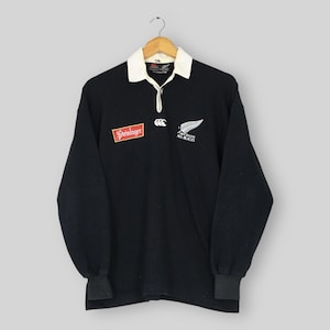 May include: Black long-sleeve rugby shirt with a white collar and cuffs. Features the Steinlager logo and the All Blacks logo with a silver fern. The shirt is hanging on a wooden hanger.