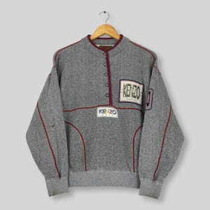 Puede incluir: Una sudadera gris de manga larga con tapeta de botones parcial, cuello alzado y ribetes de color rojo oscuro. La prenda presenta un parche de Kenzo Golf en el pecho y un parche rectangular con el logotipo de Kenzo en la parte superior derecha.
