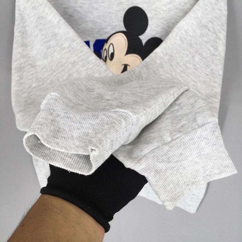 K&ouml;nnte beinhalten: Ein hellgraues Sweatshirt mit einem Mickey Mouse-Motiv. Das Motiv zeigt Mickey's Gesicht und den Buchstaben "K" in Blau. Das Sweatshirt hat gerippte B&uuml;ndchen und einen Rundhalsausschnitt.