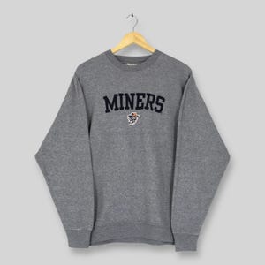 May include: A heather gray sweatshirt with the word "MINERS" in navy blue arched across the chest. Below the word "MINERS" is a small graphic of a miner. The sweatshirt is hanging on a wooden hanger.