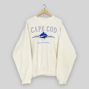 Könnte beinhalten: Cremefarbenes Sweatshirt mit dem Schriftzug "CAPE COD" in blauer Schrift über einer blau-weißen Diamantgrafik mit einer Walillustration. Das Wort "MASSACHUSETTS" ist darunter aufgedruckt.