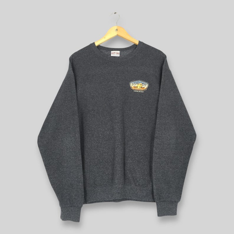 May include: A charcoal grey sweatshirt with a crew neck and long sleeves. The sweatshirt features a small, colourful patch on the upper left chest with "Funky Surf Shop" and a beach scene. It is hanging on a wooden hanger.