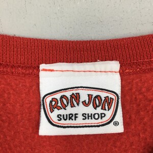May include: A close-up of a red sweatshirt collar with a white label. The label displays the "RON JON SURF SHOP" logo in red and black, with a black border. The sweatshirt is a solid, bright red.