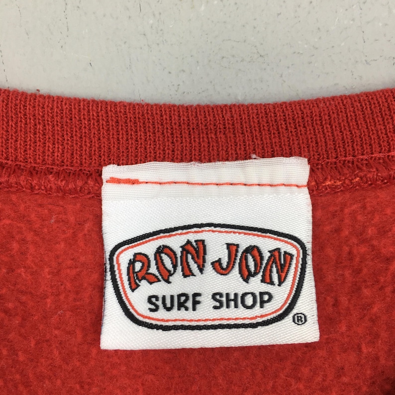 May include: A close-up of a red sweatshirt collar with a white label. The label displays the "RON JON SURF SHOP" logo in red and black, with a black border. The sweatshirt is a solid, bright red.