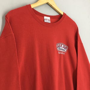 May include: A red crew neck sweatshirt featuring the Ron Jon Surfboards logo. The logo is an oval design in white and blue, with the words "Surfboards" and "Cocoa Beach" in white. The sweatshirt is suspended from a wooden hanger.
