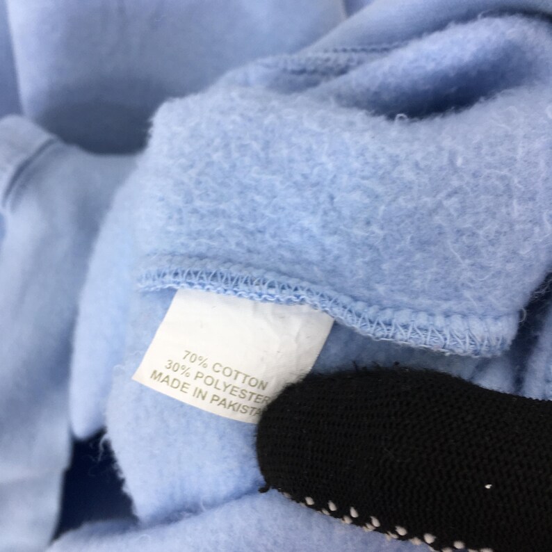 Puede incluir: Primer plano del cuello de una sudadera azul claro. Una etiqueta blanca muestra la composici&oacute;n de la tela: 70% algod&oacute;n, 30% poli&eacute;ster. La etiqueta tambi&eacute;n indica que el art&iacute;culo fue fabricado en Pakist&aacute;n. Un objeto negro es parcialmente visible.