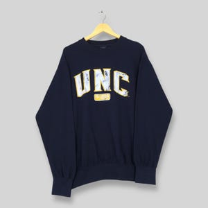Op de afbeelding: Marineblauwe sweatshirt met de letters "UNC" in wit en geel, met "Est 1889" eronder. De sweatshirt hangt aan een houten hanger.