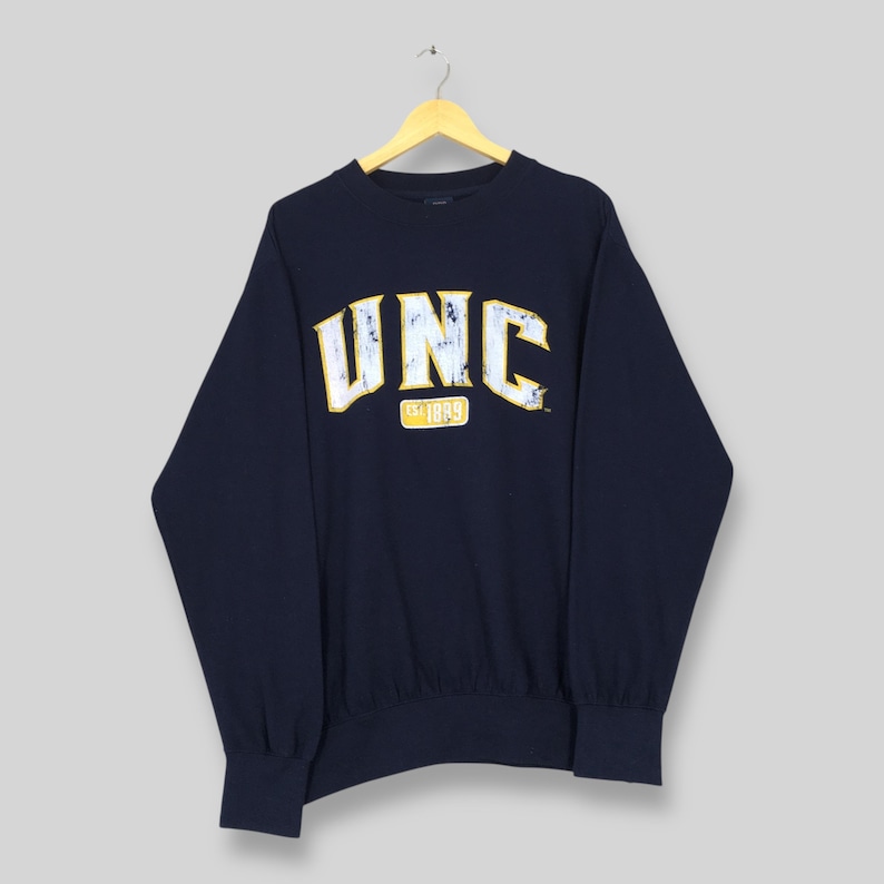 Op de afbeelding: Marineblauwe sweatshirt met de letters "UNC" in wit en geel, met "Est 1889" eronder. De sweatshirt hangt aan een houten hanger.