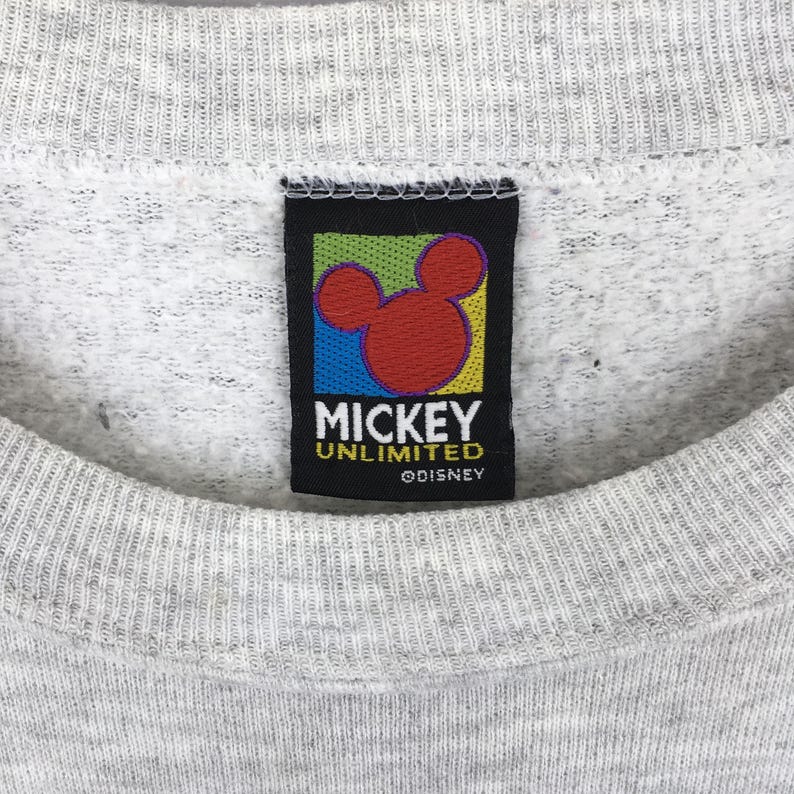 K&ouml;nnte beinhalten: Nahaufnahme eines grauen Sweatshirts mit einem schwarzen Etikett mit einem roten Mickey Mouse Kopfdesign. Das Etikett tr&auml;gt die Aufschrift "Mickey Unlimited" und "&copy;Disney" in wei&szlig;em Text. Das Sweatshirt hat einen gerippten Ausschnitt.