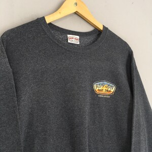 May include: Charcoal grey sweatshirt featuring the "Ron Jon Surf Shop" logo. The logo is an oval in yellow and orange, with "Cocoa Beach" below. The sweatshirt is displayed on a wooden hanger.