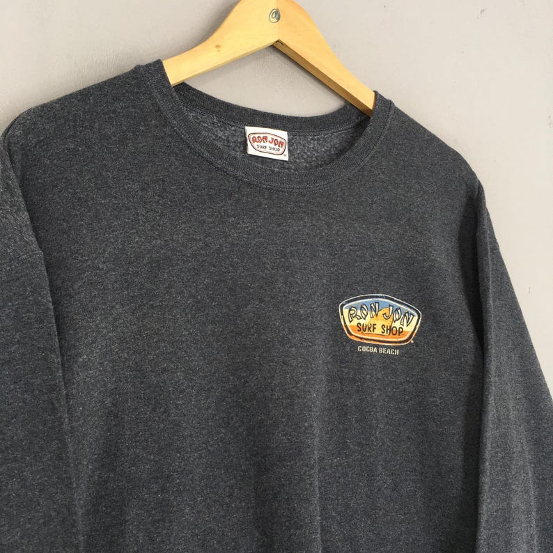 May include: Charcoal grey sweatshirt featuring the "Ron Jon Surf Shop" logo. The logo is an oval in yellow and orange, with "Cocoa Beach" below. The sweatshirt is displayed on a wooden hanger.
