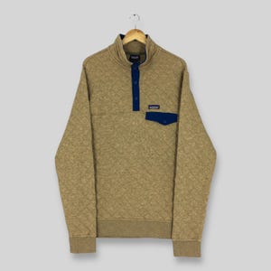 Puede incluir: Jersey acolchado color canela con cuello alzado y un pequeño bolsillo azul. El jersey tiene una tapeta abotonada y un pequeño parche azul con un logotipo. El jersey está colgado de una percha de madera.