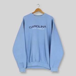 Puede incluir: Sudadera azul claro con la palabra "CAROLINA" en azul marino arqueada sobre el pecho, con "TAR HEELS" debajo. La sudadera est&aacute; colgada de una percha de madera.