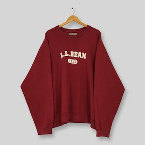 May include: A maroon sweatshirt with the text "L.L.Bean 1912" in white lettering. The sweatshirt is hanging on a wooden hanger against a light gray background. The sweatshirt has a crew neck and long sleeves.