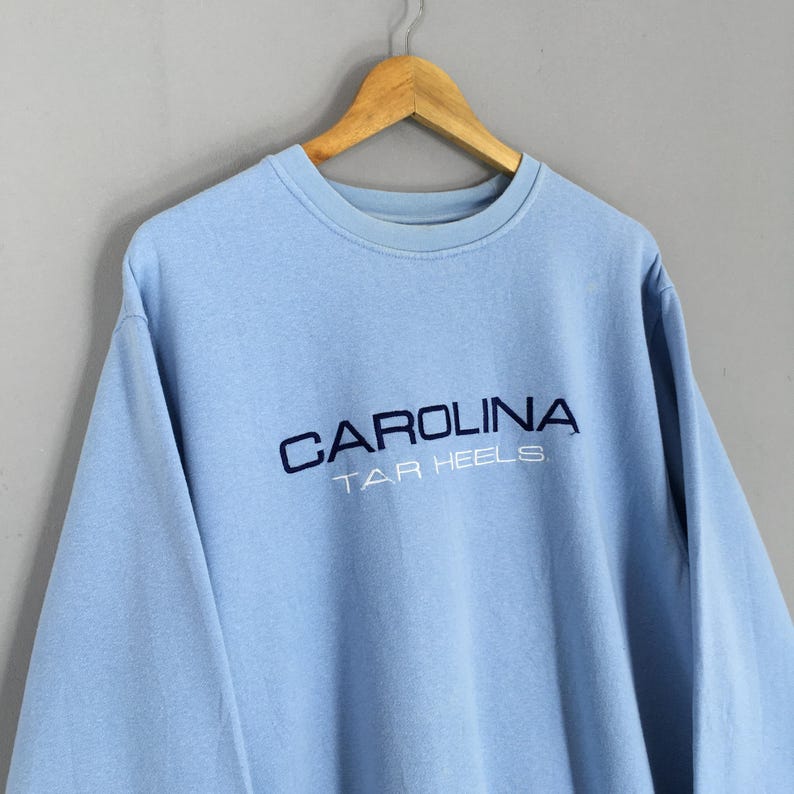 Puede incluir: Una sudadera azul claro con la palabra "CAROLINA" bordada en azul marino, con "TAR HEELS" debajo. La sudadera de cuello redondo est&aacute; colgada de una percha de madera.