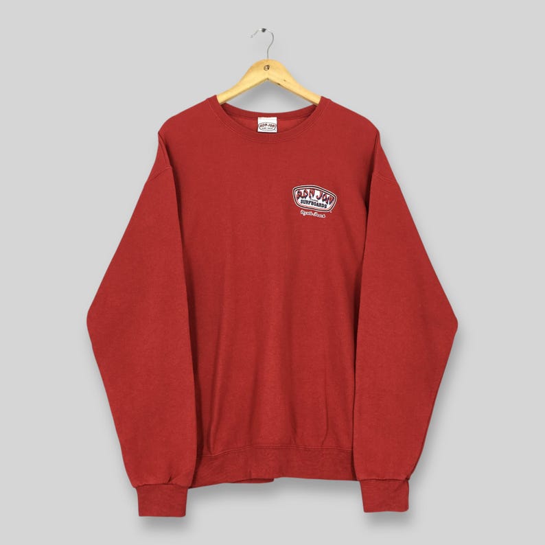 May include: A red crew neck sweatshirt featuring the Bon Jon Surfboards logo on the upper left chest. The logo is in a white and red oval shape with the words "Bon Jon Surfboards" and "Daytona Beach" in white text. The sweatshirt is hanging on a wooden hanger.