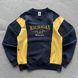 May include: A navy blue and yellow crewneck sweatshirt featuring the text MICHIGAN and Wolverines with a block M logo in the center. The garment has a front kangaroo pocket and long sleeves with color-blocked yellow side panels on the torso.