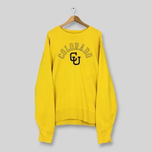 May include: A bright yellow long-sleeve crewneck sweatshirt featuring the word COLORADO arched in black lettering above a large black CU logo. The garment is displayed on a wooden hanger, showcasing a classic pullover design with ribbed cuffs.