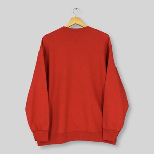 May include: A solid red sweatshirt hangs from a wooden hanger. The long-sleeved top has a crew neck and ribbed cuffs and hem. The garment is made of a soft material.