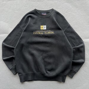 May include: A black long-sleeve crewneck sweatshirt featuring the embroidered text UCF University of Central Florida in gold and white lettering centred on the chest. The garment has a ribbed collar, cuffs, and hem with a JanSport brand label.