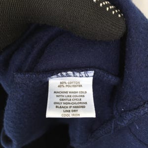 Op de afbeelding: Close-up van een marineblauw kledinglabel met wasvoorschriften. Het label luidt "60% Katoen, 40% Polyester" en bevat wasinstructies: machinewas koud met dezelfde kleuren, fijnwasprogramma, chloorvrij bleken indien nodig, aan de lijn drogen en koud strijken.