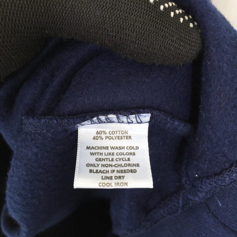 Op de afbeelding: Close-up van een marineblauw kledinglabel met wasvoorschriften. Het label luidt "60% Katoen, 40% Polyester" en bevat wasinstructies: machinewas koud met dezelfde kleuren, fijnwasprogramma, chloorvrij bleken indien nodig, aan de lijn drogen en koud strijken.