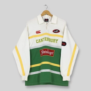 May include: A long-sleeved rugby shirt in white, green, and yellow, with the word "Canterbury" across the chest. The shirt features the "Steinlager" logo and a small "CNZ Rugby" patch. The sleeves have yellow stripes.
