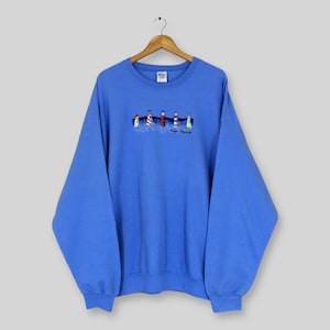 Può includere: Una felpa blu con ricami di fari e la scritta "Door County". I fari sono di vari colori, tra cui rosso, bianco e blu. La felpa è appesa a una gruccia di legno.