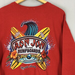 May include: Red sweatshirt with a graphic of surfboards, a wave, and the text "Ron Jon Surfboards." The design includes hibiscus flowers and the words "Myrtle Beach." The sweatshirt has a crew neck and long sleeves.