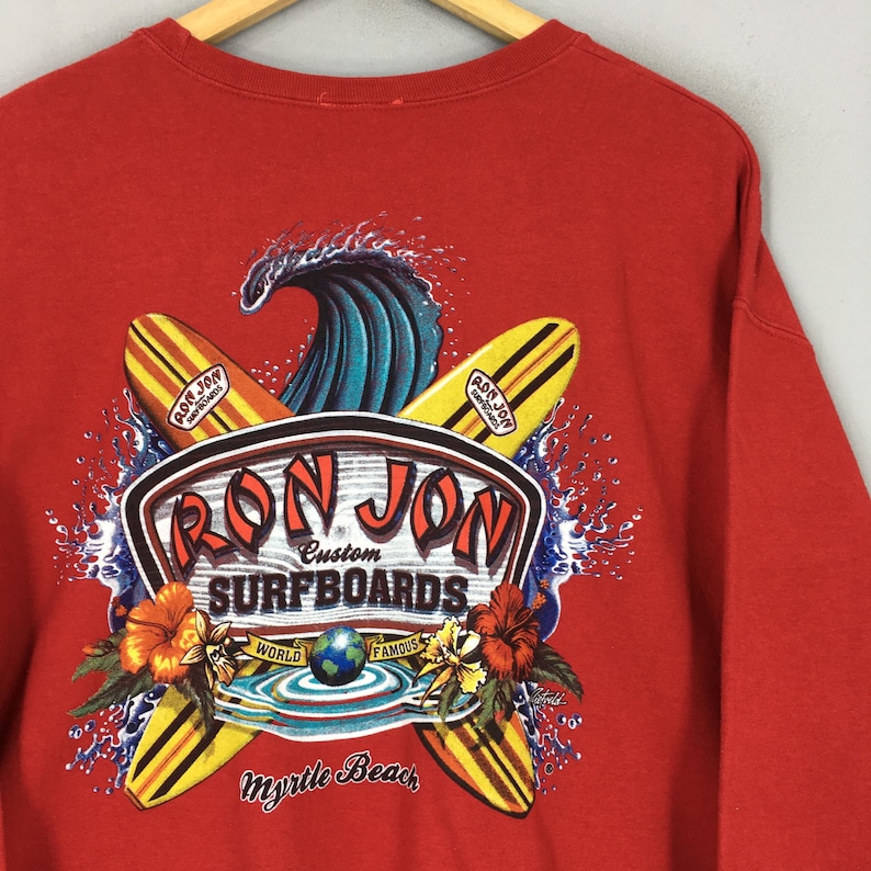 May include: Red sweatshirt with a graphic of surfboards, a wave, and the text "Ron Jon Surfboards." The design includes hibiscus flowers and the words "Myrtle Beach." The sweatshirt has a crew neck and long sleeves.