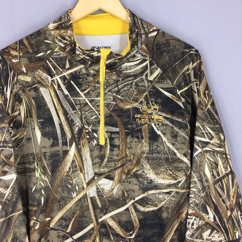 Puede incluir: Camisa de manga larga de camuflaje con cuello y cremallera amarillos. La camisa presenta un logotipo de Realtree Max5 y un patr&oacute;n de follaje marr&oacute;n, verde y beige. La camisa est&aacute; colgada en una percha de madera.