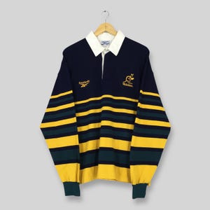 Könnte beinhalten: Marineblau-gelb gestreiftes Rugby-Shirt mit weißem Kragen. Es hat das Reebok-Logo und ein Känguru-Emblem mit dem Text "Wallabies". Das Shirt mit langen Ärmeln hängt an einem Holzkleiderbügel.